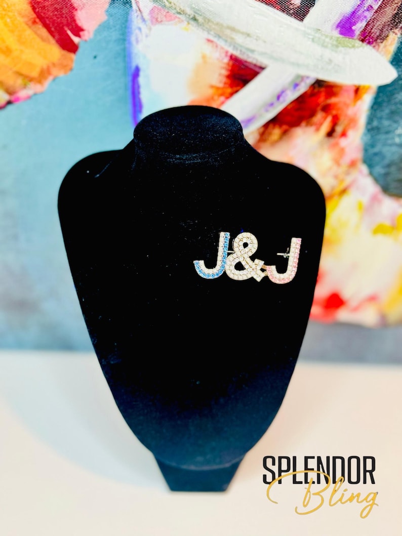 Pu&ograve; includere: Spilla color argento con le lettere "J & J" in blu e rosa, impreziosita da cristalli trasparenti. La spilla &egrave; esposta su un busto di velluto nero. Il testo "SPLENDOR Bling" &egrave; visibile in basso.