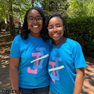 May include: Two people wearing matching blue t-shirts with a pink, blue, and white graphic design. The graphic design features the letters "JJ" with a white line across the top.