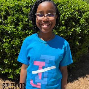 May include: A young person wearing a blue t-shirt with a pink, blue, and white graphic design. The graphic design features a stylized letter "J" with a diagonal line through it.