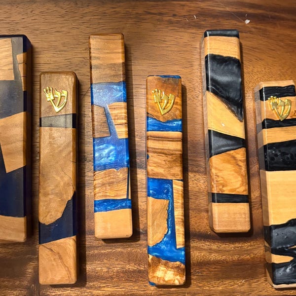 May include: A collection of six wooden mezuzah cases with varying designs. The cases feature natural wood tones, dark blue, and black resin accents. Some have a gold-colored emblem.