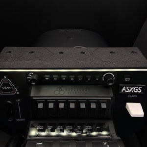 May include: Black control panel with a white light strip, labelled "ASXGGS FLAPS" and "AUTO PILOT". The panel has a row of black toggle switches with white labels, including "MASTER WARNING", "ENGINE FIRE", "LOW FUEL PRESSURE", "LOW FUEL", "AMP", "STARTER ENGAGED", "APU", "PARKING BRAKE", "LOW VOLTS", and "DOOR".