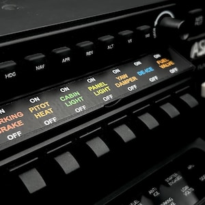 Könnte beinhalten: Nahaufnahme eines schwarzen Flugzeug-Bedienfelds mit verschiedenen beleuchteten Schaltern und Beschriftungen. Das Panel enthält Bedienelemente für Parkbremse, Staurohrheizung, Kabinenlicht und Enteisung. Rechts befindet sich ein kleines weißes rechteckiges Objekt.