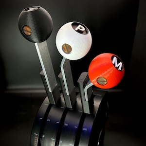 May include: A close-up of a black, white, and red gear shift assembly. The gear shift knobs are spherical, with the letters "P" and "M" on the white and red knobs. The assembly has a black base with silver levers.
