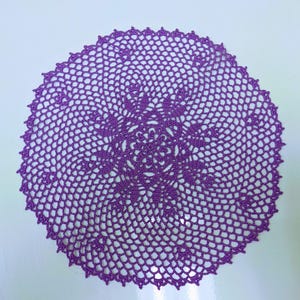 May include: A handmade, circular doily in a vibrant purple hue. The crocheted piece features a detailed, openwork design with a central floral motif and a scalloped edge. This decorative item is suitable for home decor.