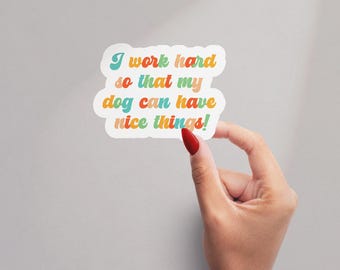 May include: A white sticker with the text "I work hard so that my dog can have nice things!" in colorful retro font. The sticker is held by a hand with a red painted fingernail.