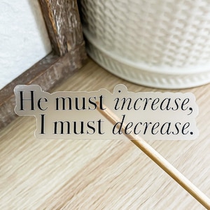 May include: A clear sticker with the text "He must increase, I must decrease." on a wooden surface.