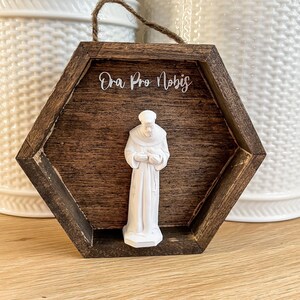 Op de afbeelding: Een zeshoekig houten frame met een donkerbruine vlek. Het frame heeft een wit opschrift met de tekst "Ora Pro Nobis". Binnen het frame bevindt zich een wit beeldje van een heilige.