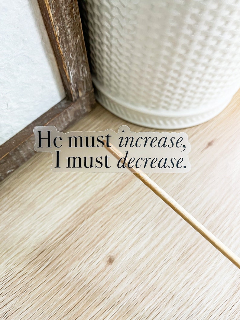 May include: A clear sticker with the text "He must increase, I must decrease." on a wooden surface.