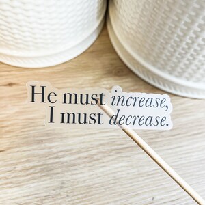 Może przedstawiać: Przezroczysta naklejka z tekstem "He must increase, I must decrease." na drewnianej powierzchni.