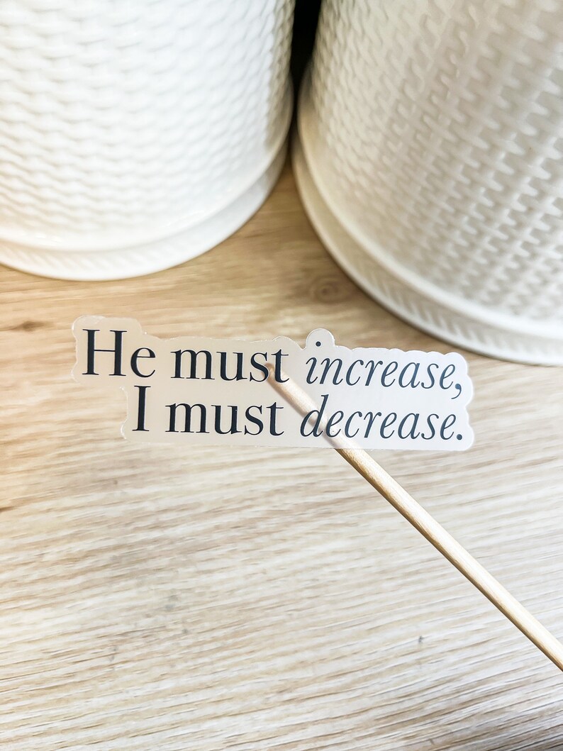 Może przedstawiać: Przezroczysta naklejka z tekstem "He must increase, I must decrease." na drewnianej powierzchni.