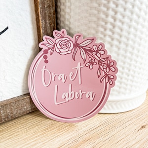 May include: A pink round sticker with a floral design and the text "Ora et Labora" in a darker pink.