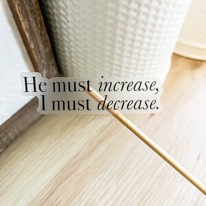 Może przedstawiać: Przezroczysta naklejka z tekstem "He must increase, I must decrease." na drewnianej powierzchni.