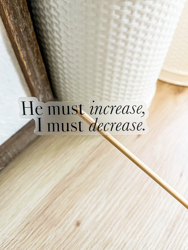 Może przedstawiać: Przezroczysta naklejka z tekstem "He must increase, I must decrease." na drewnianej powierzchni.