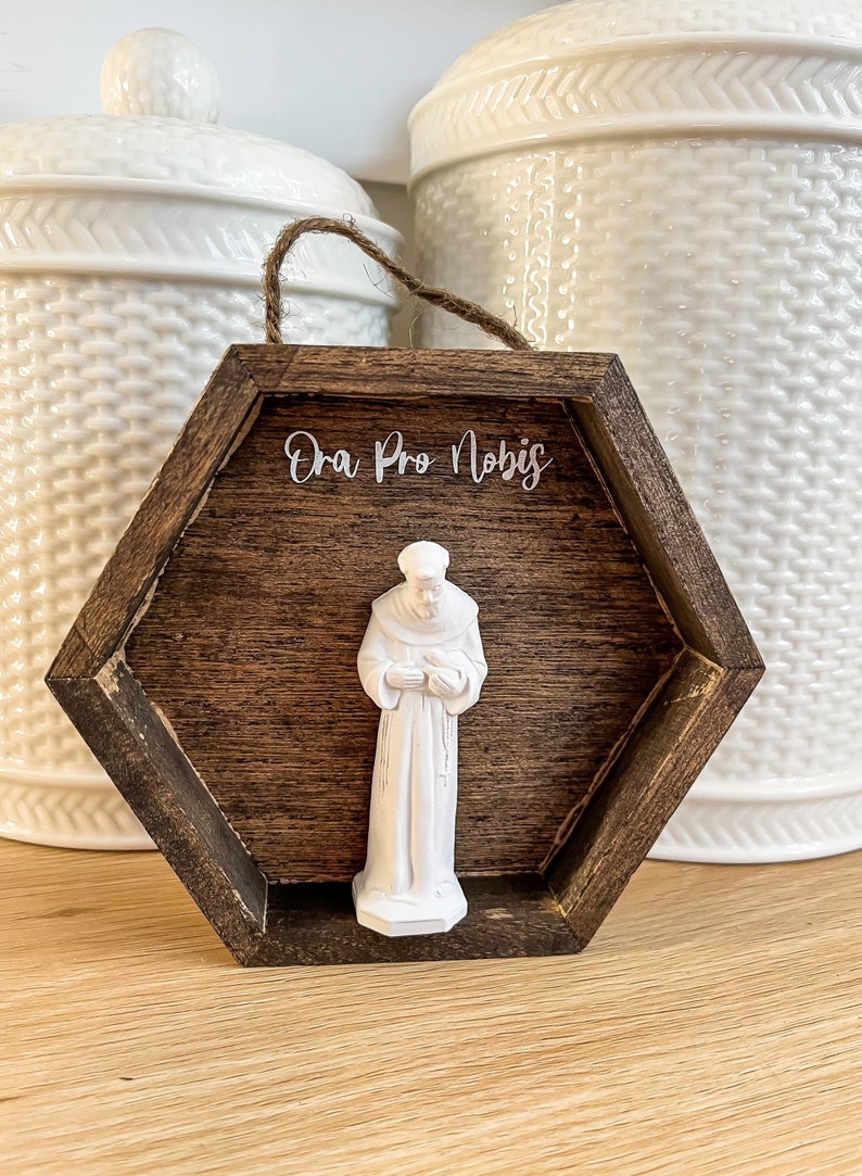 Op de afbeelding: Een zeshoekig houten frame met een donkerbruine vlek. Het frame heeft een wit opschrift met de tekst "Ora Pro Nobis". Binnen het frame bevindt zich een wit beeldje van een heilige.