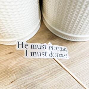 Może przedstawiać: Biała naklejka z tekstem "He must increase, I must decrease." na drewnianej powierzchni.