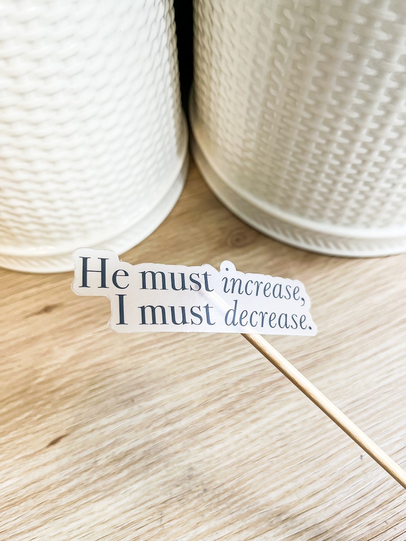 Może przedstawiać: Biała naklejka z tekstem "He must increase, I must decrease." na drewnianej powierzchni.