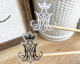 Op de afbeelding: Twee transparante stickers met een gestileerde letter M bekroond met een kroon. De ene sticker toont het ontwerp in effen wit, terwijl de andere sticker hetzelfde ontwerp in effen zwart toont. Elke sticker is bevestigd aan een dun houten stokje.