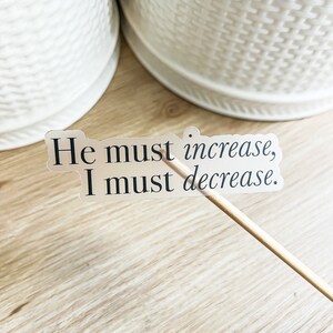 Może przedstawiać: Przezroczysta naklejka z tekstem "He must increase, I must decrease." na drewnianej powierzchni.