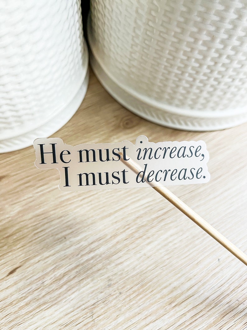 Może przedstawiać: Przezroczysta naklejka z tekstem "He must increase, I must decrease." na drewnianej powierzchni.