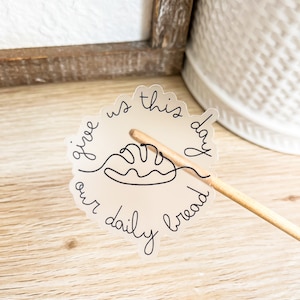 May include: A clear sticker with the text "Give us this day our daily bread" and a simple line drawing of a loaf of bread.