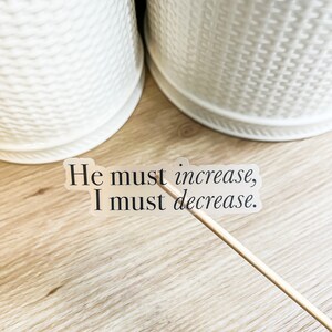 Może przedstawiać: Przezroczysta naklejka z tekstem "He must increase, I must decrease." na drewnianej powierzchni.