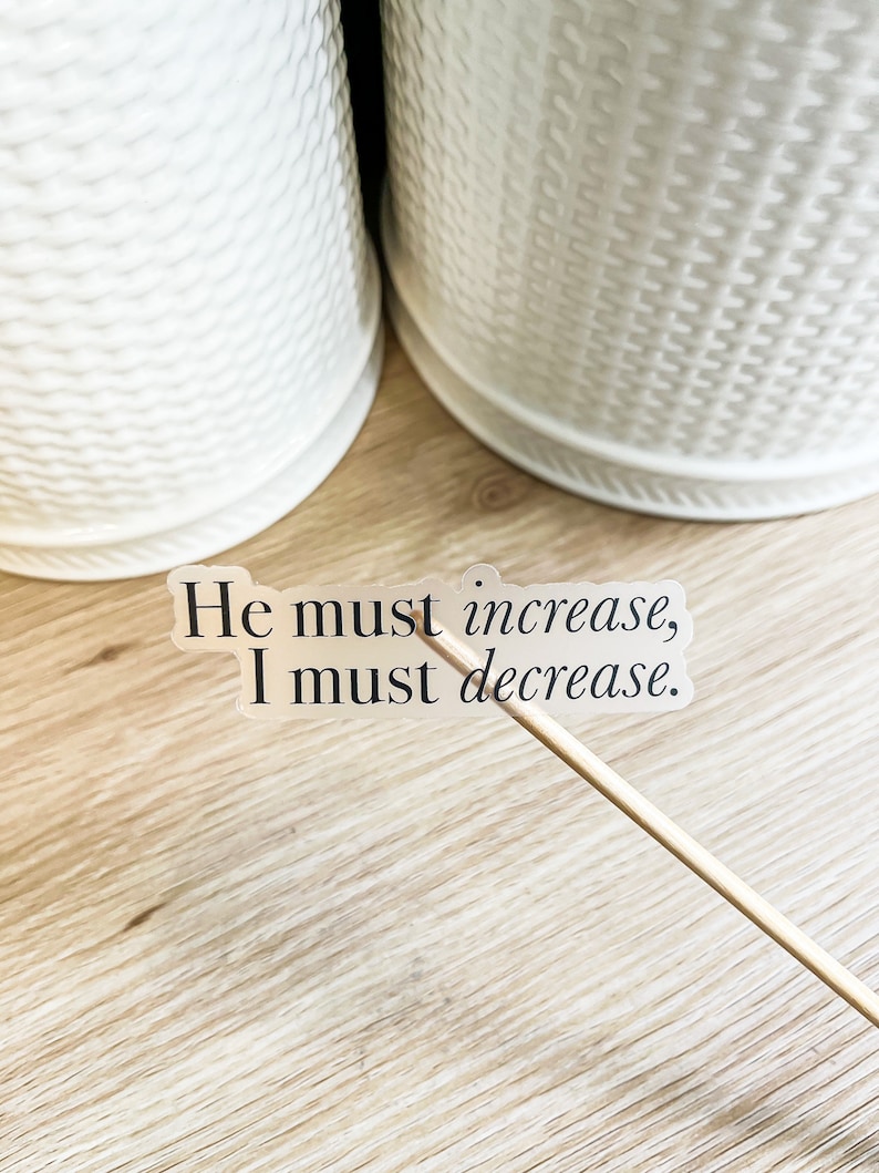 Może przedstawiać: Przezroczysta naklejka z tekstem "He must increase, I must decrease." na drewnianej powierzchni.