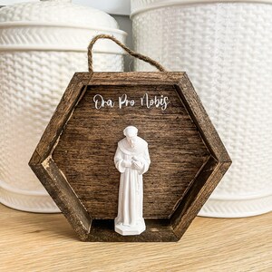 Op de afbeelding: Een zeshoekig houten frame met een wit beeldje van een heilige erin. Het frame heeft de woorden "Ora Pro Nobis" erop geschreven.