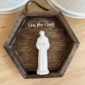 Op de afbeelding: Een wit beeldje van een heilige in een bruin zeshoekig houten frame met de tekst "Ora Pro Nobis" in wit.