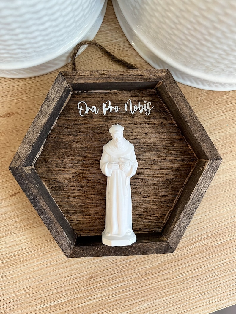 Op de afbeelding: Een wit beeldje van een heilige in een bruin zeshoekig houten frame met de tekst "Ora Pro Nobis" in wit.