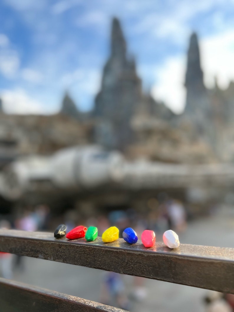 K&ouml;nnte beinhalten: Eine Reihe von sechs kleinen, bunten, tropfenf&ouml;rmigen Objekten aus Kunststoff auf einem Metallgel&auml;nder. Die Objekte sind schwarz, rot, gr&uuml;n, gelb, blau und wei&szlig;.