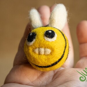 Könnte beinhalten: Eine gelbe Filz-Biene mit schwarzen Streifen und weißen Filzflügeln. Die Biene hat ein lächelndes Gesicht mit schwarzen Augen und weißen Zähnen.