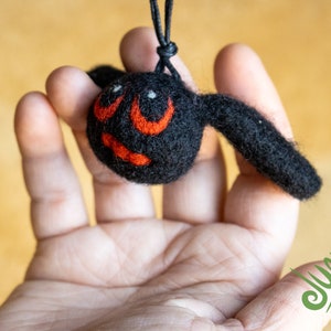 Könnte beinhalten: Eine schwarze Filzfledermaus mit roten Augen und einem schwarzen Filzkörper. Die Fledermaus hängt an einer schwarzen Schnur und wird in der Hand einer Person gehalten.
