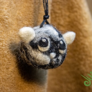 Op de afbeelding: Een zwart-witte vilten dierenornament met een schattig gezicht, hangend aan een zwart koord. Het ornament is gevormd als een lemur met grote ogen en een kleine neus.