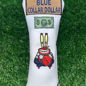 Może przedstawiać: Biały pokrowiec na główkę kija golfowego z napisem "BLUE COLLAR DOLLAR" i kreskówkowym wizerunkiem Pana Kraba. Pokrowiec ma czarno-biały pasiasty spód i zieloną grafikę banknotu.