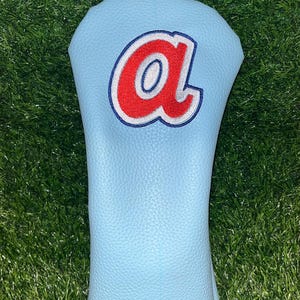 Könnte beinhalten: Hellblaue Golfschlägerhaube mit einem rot-weißen Buchstaben "a"-Design. Die Hülle hat eine rote Umrandung und ist vor einem Hintergrund aus grünem Gras platziert.