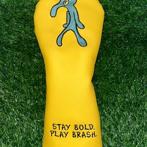 Może przedstawiać: Żółta osłona na główkę kija golfowego z turkusową postacią z kreskówki i napisem "STAY BOLD. PLAY BRASH." Pokrowiec wykonany jest z gładkiego materiału i ma chronić kije golfowe.