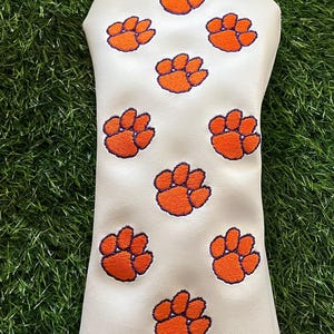 May include: A white golf club headcover featuring a repeating pattern of embroidered orange paw prints with purple outlines. The headcover has a smooth white surface and a solid purple fabric trim along the bottom edge, designed for golf clubs.