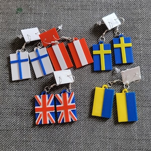 May include: A selection of flag earrings, featuring designs from Finland, Austria, Sweden, the UK, and Ukraine. The earrings are rectangular with silver hooks and white backing.