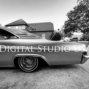 以下が含まれることがあります： モノクロ写真のクラシックなローライダー車が私道に駐車されています。銀色の車は、複雑なクロムワイヤーホイールを備えています。「DIGITAL STUDIO USA」というテキストが車の側面に重ねられています。背景には家と木があります。