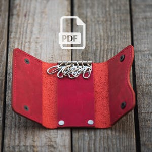 Könnte beinhalten: Roter Leder-Schlüsselanhänger mit einem Schlüsselring aus Metall und fünf Schlüsselschlaufen. Der Schlüsselanhänger ist geöffnet und der Schlüsselring ist sichtbar. Der Hintergrund ist eine Holzoberfläche.