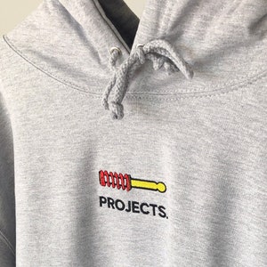 Op de afbeelding: Een grijs hoodie met een klein geborduurd logo van een gele en rode veer met het woord "PROJECTS." eronder.
