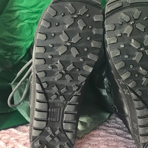 Puede incluir: Primer plano de las suelas de goma negra de un par de botas negras. Las suelas tienen un patr&oacute;n de banda de rodadura en forma de diamante.