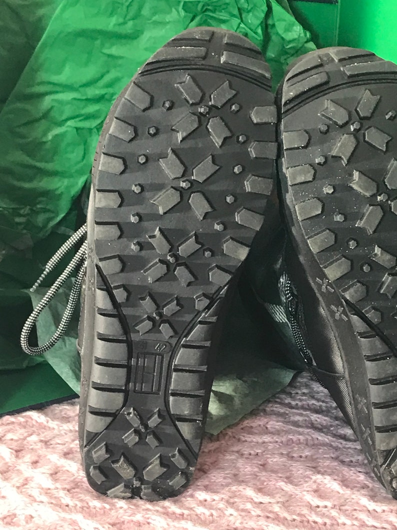 Puede incluir: Primer plano de las suelas de goma negra de un par de botas negras. Las suelas tienen un patr&oacute;n de banda de rodadura en forma de diamante.