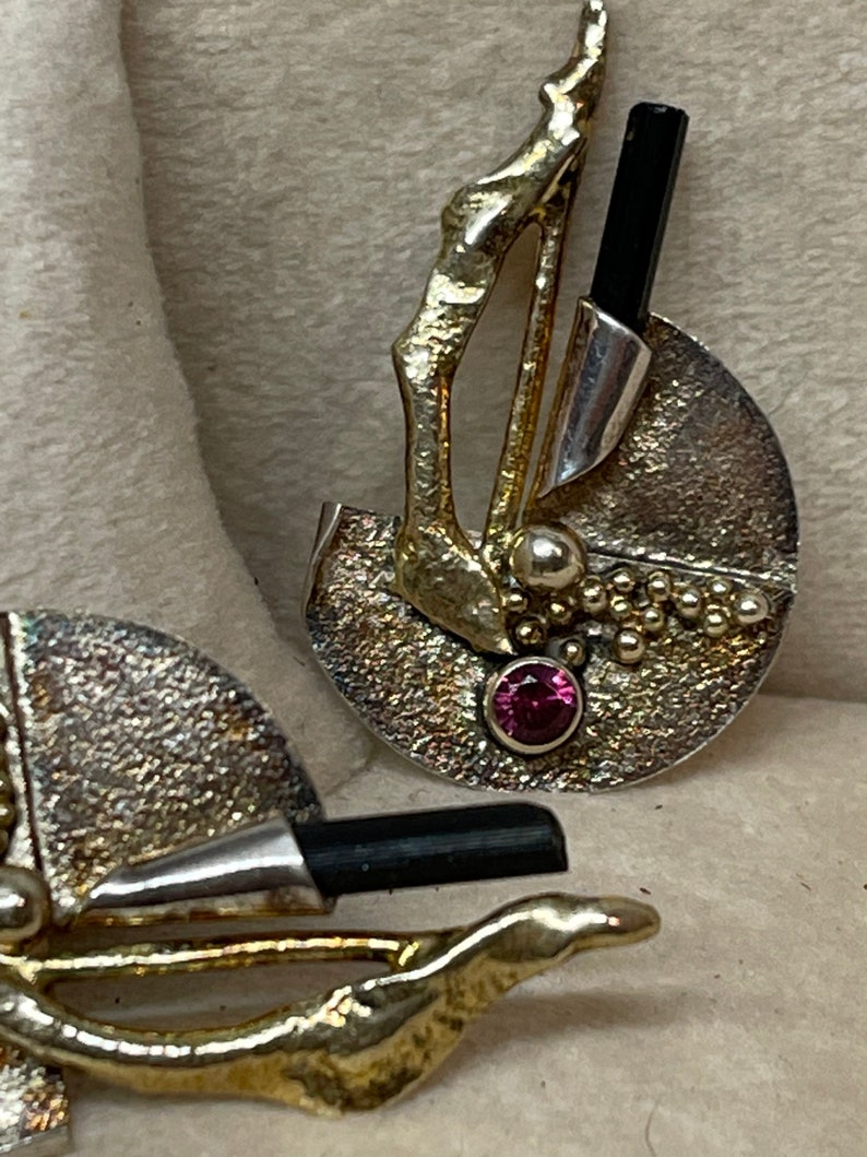 May include: Artisan-crafted earrings featuring a textured silver-tone disc with gold-tone accents. Each earring has a black rectangular stone, a small pink gemstone, and a cluster of tiny gold-tone beads. The design is abstract and unique.