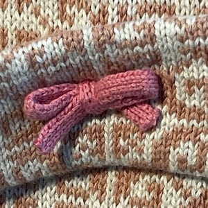 May include: Close-up of a knitted fabric with a pink bow. The fabric features a pattern of alternating white and peach-coloured stripes. The bow is a solid pink colour and is attached to the fabric.