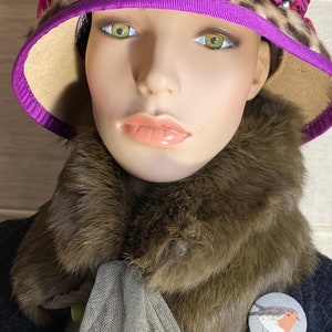 May include: A brown faux fur collar with a light brown scarf and a fabric pin with a robin illustration. The collar is worn over a dark blue jacket. The hat is a leopard print with a purple band and a brown brim.