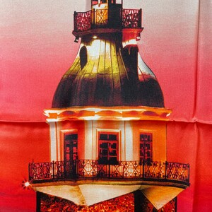 Puede incluir: Una torre alta y blanca con un techo rojo y ventanas negras. La torre tiene una serie de balcones con barandillas negras. La torre está iluminada por la noche, y el fondo es una puesta de sol roja y naranja.