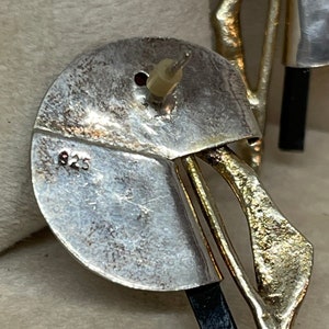 May include: Close-up of a silver earring with a post back. The earring has a circular design with a section cut out, and a gold-colored, abstract shape. The number "925" is stamped on the silver.