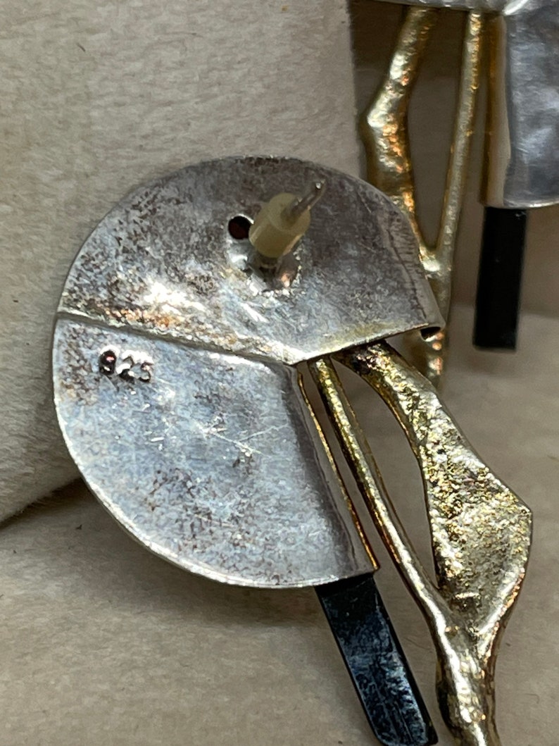 May include: Close-up of a silver earring with a post back. The earring has a circular design with a section cut out, and a gold-colored, abstract shape. The number "925" is stamped on the silver.