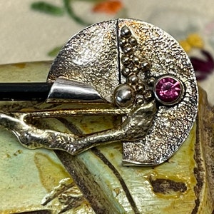 May include: A silver-toned brooch with a textured, semi-circular design. It features a black rectangular stone, a cluster of small silver spheres, and a round, pink gemstone. The brooch has a branch-like element extending from the base.